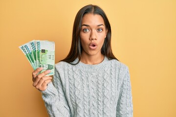 Beautiful hispanic woman holding 10000 south korean won banknotes scared and amazed with open mouth for surprise, disbelief face