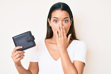 Young beautiful latin girl holding leather wallet covering mouth with hand, shocked and afraid for...