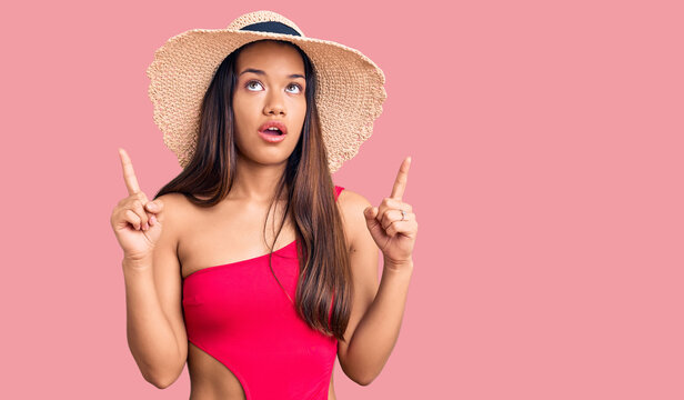 Young beautiful latin girl wearing swimwear and summer hat amazed and surprised looking up and pointing with fingers and raised arms.
