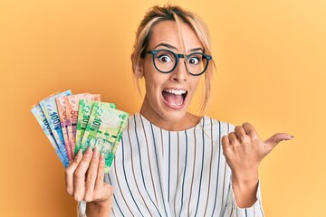 Beautiful blonde woman holding south african rand banknotes pointing thumb up to the side smiling...