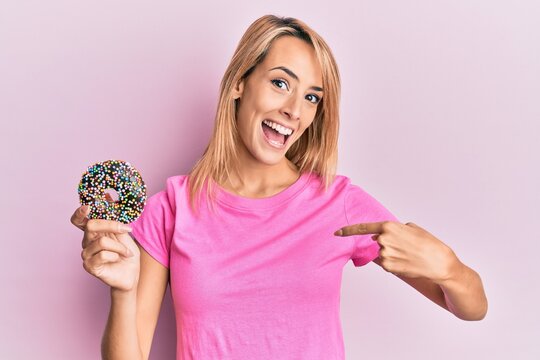 Beautiful blonde woman holding donut pointing finger to one self smiling happy and proud