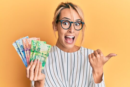 Beautiful blonde woman holding south african rand banknotes pointing thumb up to the side smiling happy with open mouth