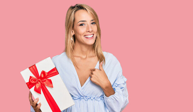 Beautiful blonde woman holding gift pointing finger to one self smiling happy and proud