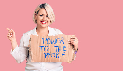 Young blonde plus size woman holding power to the people banner smiling happy pointing with hand and finger to the side