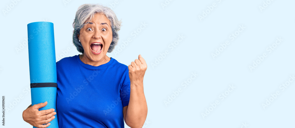 Poster Senior woman with gray hair holding yoga mat screaming proud, celebrating victory and success very excited with raised arms