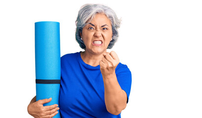 Senior woman with gray hair holding yoga mat annoyed and frustrated shouting with anger, yelling crazy with anger and hand raised