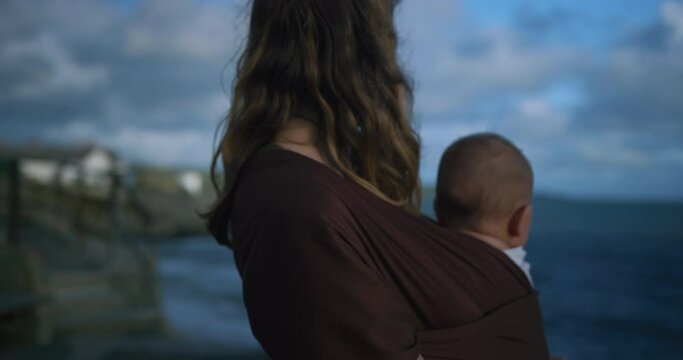 A young mother is standing on the coast with her baby in a sling in the autumn