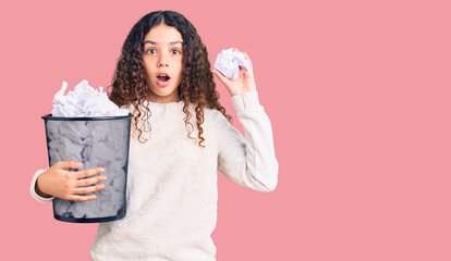 Beautiful kid girl with curly hair holding paper bin full of crumpled papers scared and amazed with...