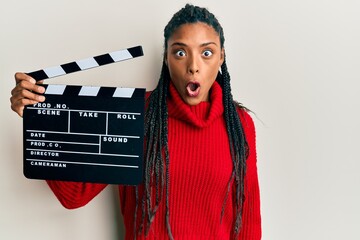 African american woman with braids holding video film clapboard scared and amazed with open mouth...