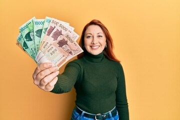 Beautiful redhead woman holding south korean won banknotes looking positive and happy standing and smiling with a confident smile showing teeth