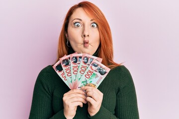 Beautiful redhead woman holding 100 new zealand dollars banknote making fish face with mouth and squinting eyes, crazy and comical.