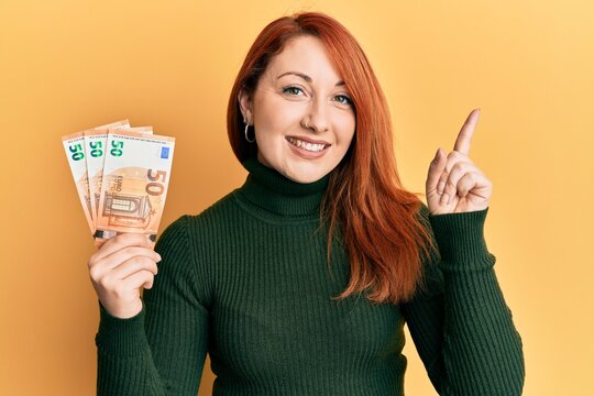 Beautiful redhead woman holding bunch of 50 euro banknotes smiling happy pointing with hand and finger to the side