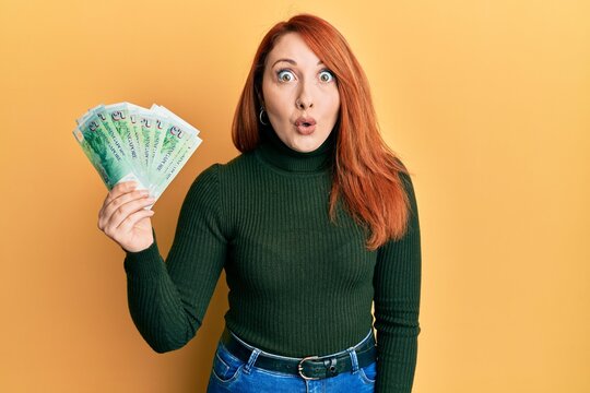 Beautiful redhead woman holding 5 singapore dollars banknotes scared and amazed with open mouth for surprise, disbelief face
