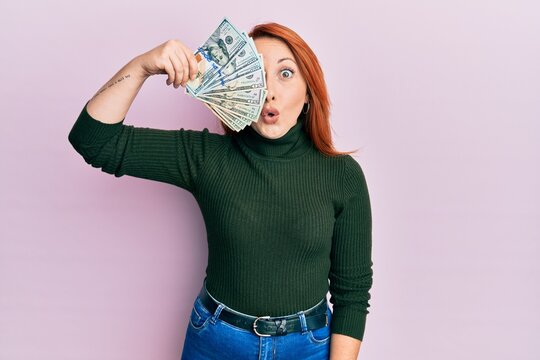 Beautiful redhead woman holding dollars banknote over eye scared and amazed with open mouth for surprise, disbelief face