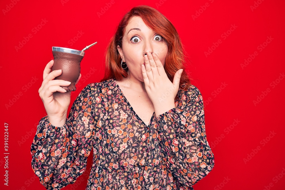 Wall mural Young beautiful redhead woman drinking mate infusion beverage over isolated red background covering mouth with hand, shocked and afraid for mistake. Surprised expression