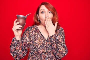 Young beautiful redhead woman drinking mate infusion beverage over isolated red background covering...