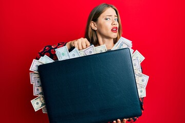 Beautiful young blonde woman holding briefcase full of dollars clueless and confused expression. doubt concept.