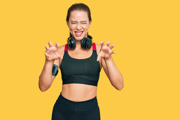 Beautiful blonde woman wearing gym clothes and using headphones smiling funny doing claw gesture as...