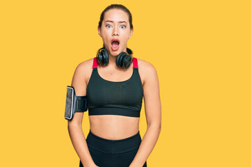 Beautiful blonde woman wearing gym clothes and using headphones afraid and shocked with surprise and amazed expression, fear and excited face.