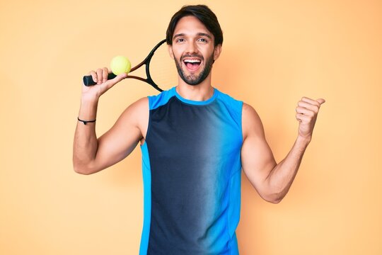 Handsome hispanic man playing tennis holding racket and ball pointing thumb up to the side smiling happy with open mouth