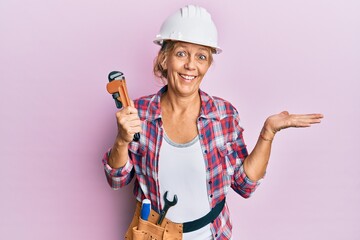 Middle age blonde woman wearing hardhat holding a wrench celebrating achievement with happy smile and winner expression with raised hand