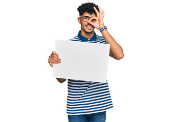 Young arab man holding blank empty banner smiling happy doing ok sign with hand on eye looking through fingers