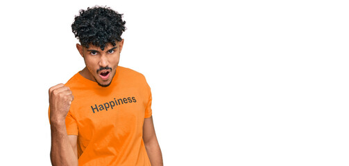Young arab man wearing tshirt with happiness word message angry and mad raising fist frustrated and furious while shouting with anger. rage and aggressive concept.