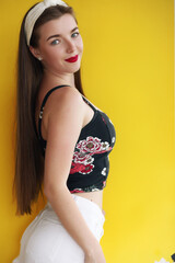Happy girl in vintage denim shorts standing in confident pose on yellow background. Indoor portrait of enchanting young lady in summer casual outfit plays with her hair.