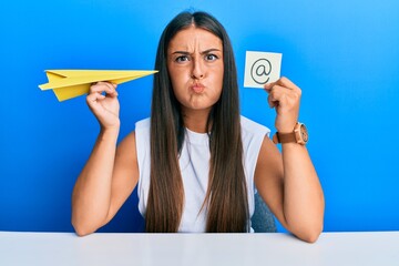 Beautiful hispanic woman holding paper plane and email symbol puffing cheeks with funny face. mouth...