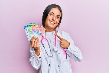 Beautiful hispanic woman wearing medical uniform holding swiss franc banknotes smiling happy pointing with hand and finger