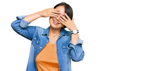 Young beautiful hispanic woman with short hair wearing casual denim jacket covering eyes with hands smiling cheerful and funny. blind concept.