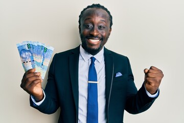 Handsome young black man wearing business suit holding 100 rands banknotes screaming proud,...