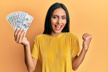 Young brunette woman holding dollars pointing thumb up to the side smiling happy with open mouth