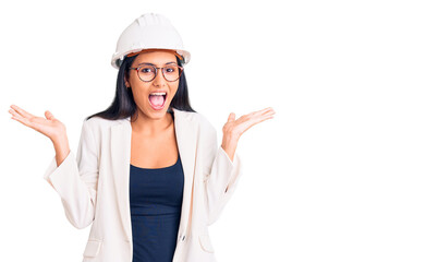 Young beautiful latin girl wearing architect hardhat and glasses celebrating crazy and amazed for success with arms raised and open eyes screaming excited. winner concept