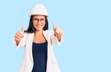 Young beautiful latin girl wearing architect hardhat and glasses approving doing positive gesture with hand, thumbs up smiling and happy for success. winner gesture.