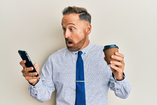Handsome middle age business man using smartphone and drinking a cup of coffee afraid and shocked with surprise and amazed expression, fear and excited face.