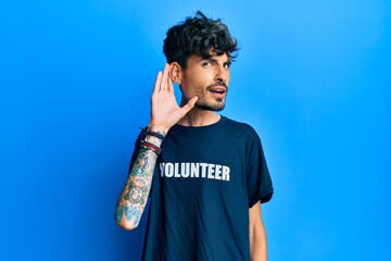 Young hispanic man wearing volunteer t shirt smiling with hand over ear listening an hearing to...