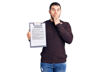 Young handsome man holding clipboard with terms and conditions document covering mouth with hand,...
