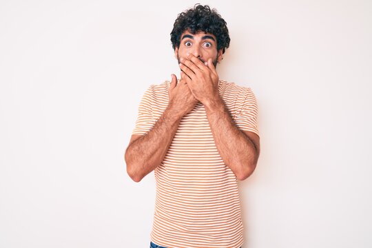 Handsome young man with curly hair and bear wearing casual striped tshirt shocked covering mouth with hands for mistake. secret concept.