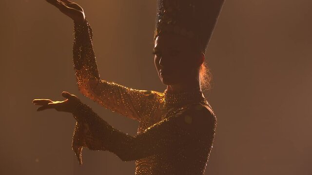 Theatrical dance show. Actress, dressed as the queen of ancient Egypt, dances in a dark studio. Silhouette of slender women on a smoky background with backlight. Close up.