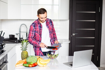 It's so delicious. Casual happy young man preparing salad at home in loft kitchen and smiling, using laptop.
