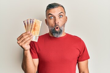 Middle age handsome man holding 500 norwegian krone banknotes scared and amazed with open mouth for...
