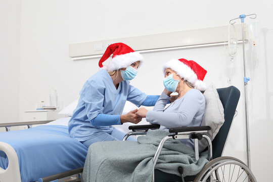 Christmas day isolated in hospital room with coronavirus, nurse take comfort elderly woman celebrating xmas wearing red caps and surgical protective medical masks