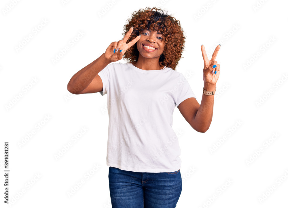 Wall mural Young african american woman wearing casual white tshirt smiling looking to the camera showing fingers doing victory sign. number two.