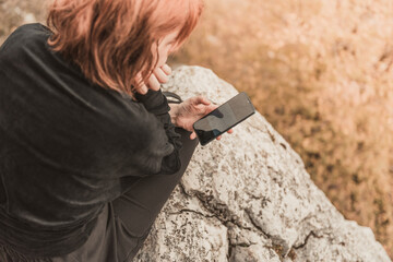 The red-haired traveler girl writes SMS on her smartphone sitting on the mountain rock. The concept of travel, technology.