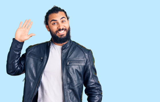 Young arab man wearing casual leather jacket waiving saying hello happy and smiling, friendly welcome gesture