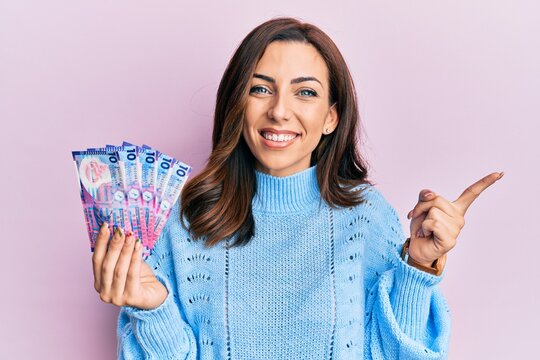 Young brunette woman holding hong kong 10 dollars banknotes smiling happy pointing with hand and finger to the side