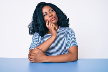 Beautiful african woman wearing casual clothes sitting on the table serious face thinking about...