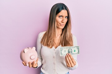 Brunette young woman holding one dollar banknote and piggy bank clueless and confused expression. doubt concept.