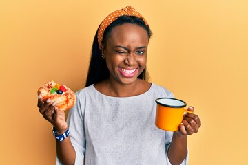 Young african woman drinking coffee and eating pastry winking looking at the camera with sexy...